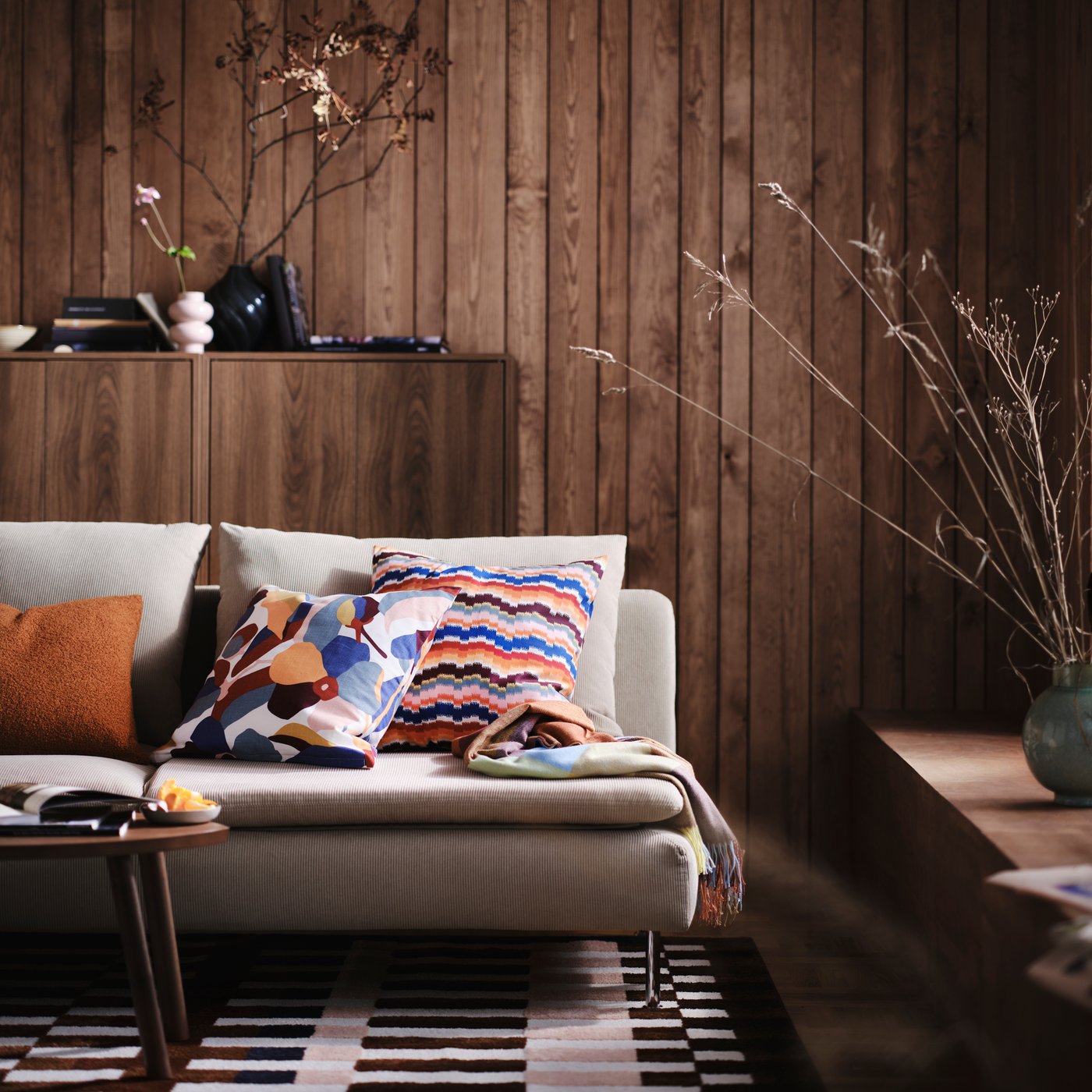A multicoloured VIDEOXBÄR cushion cover with abstract floral patterns is on a beige sofa in a wood-panelled living room.