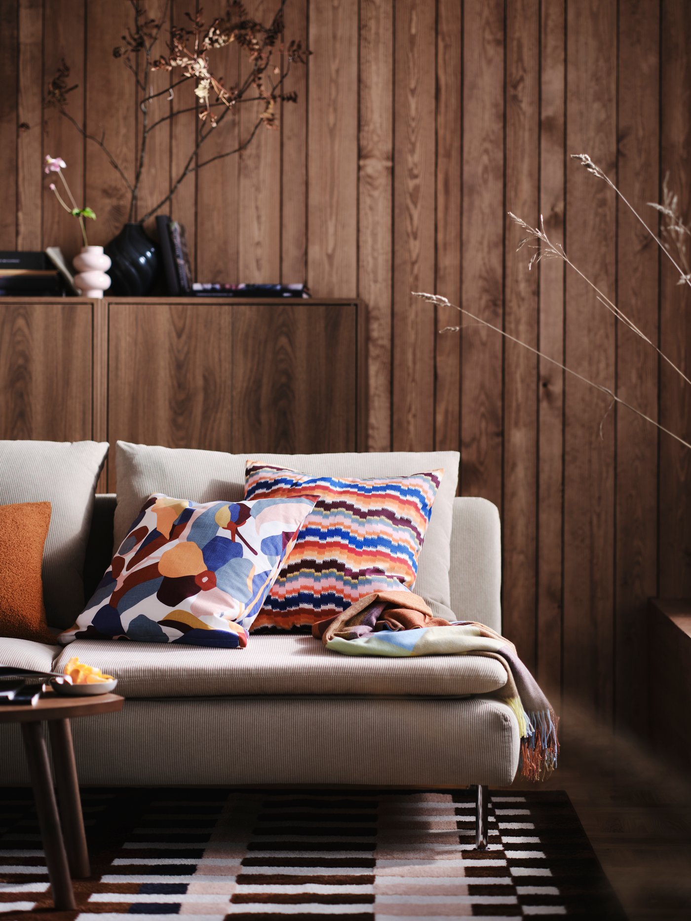 Une housse de coussin VIDEOXBÄR multicolore à motifs fleuris abstraits revêtant un coussin posé sur un canapé beige, dans un salon lambrissé de bois.