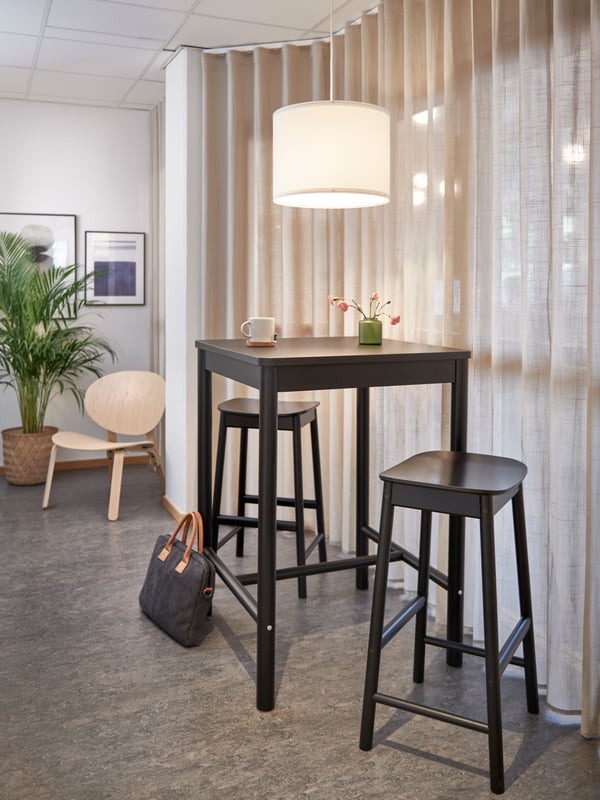 Rum med ett högt svart bord och två matchande barstolar. På bordet står en vit mugg och en liten grön vas med blommor, medan en stor vit taklampa hänger ovanför. I bakgrunden syns en trästol intill en krukväxt, några inramade bilder på väggen och en svart väska med bruna handtag på golvet. Rummet ramas in av beige gardiner och ett grått golv.