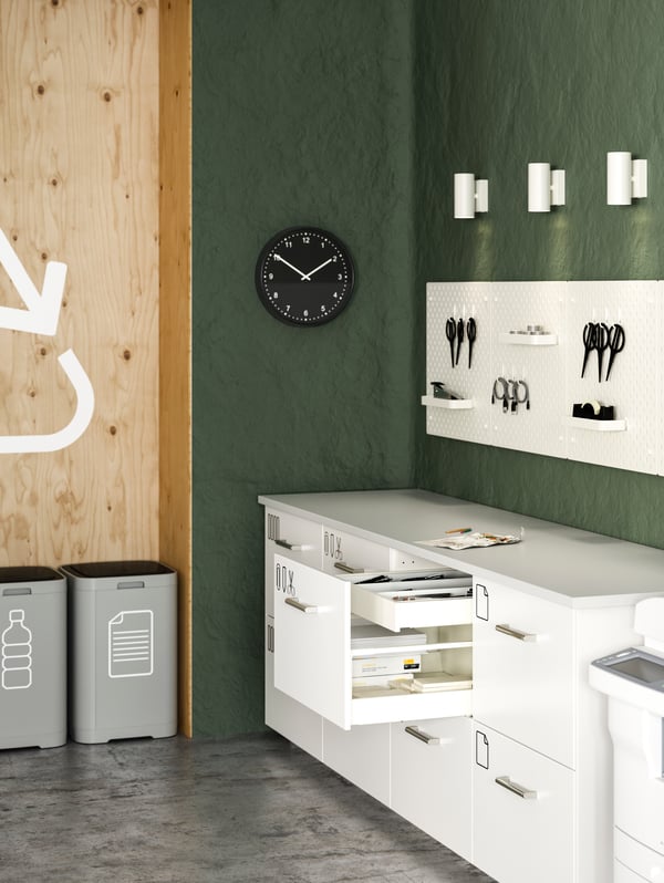 A copy room with several white drawers, white pegboards, white spotlights, waste sorting bins and a black wall clock.
