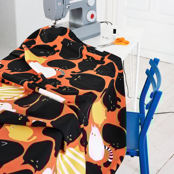 På ett bord vid en grå symaskin ligger ett orange tyg med möster av svarta katter. 