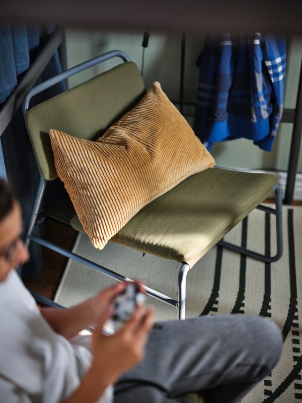 A person with a game controller sitting next to an olive green LINNEBÄCK easy chair with a cushion in a ÅSVEIG cushion cover.