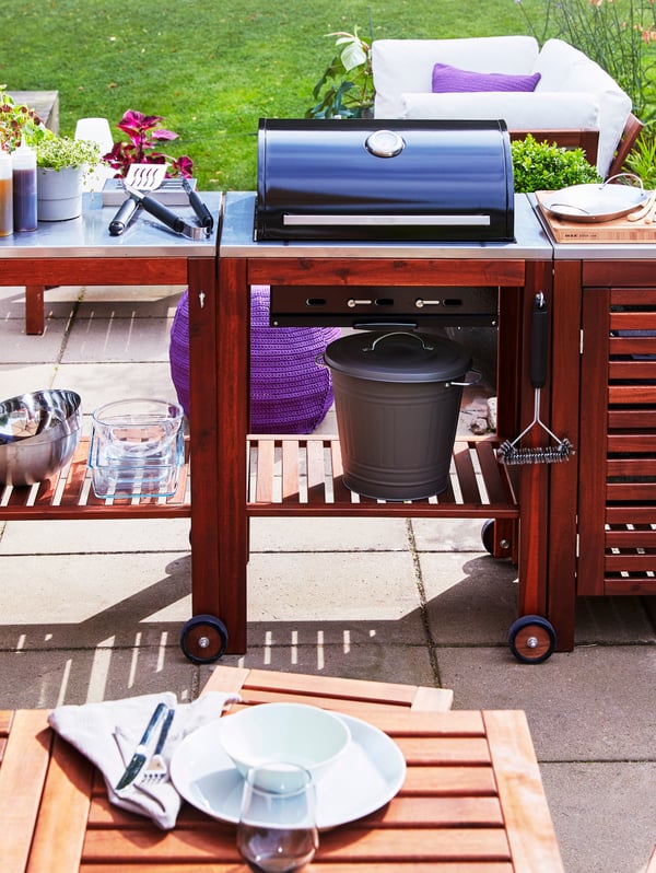 A charcoal barbecue with trolley and cabinet in acacia wood stands on a patio in front of an outdoor dining table.