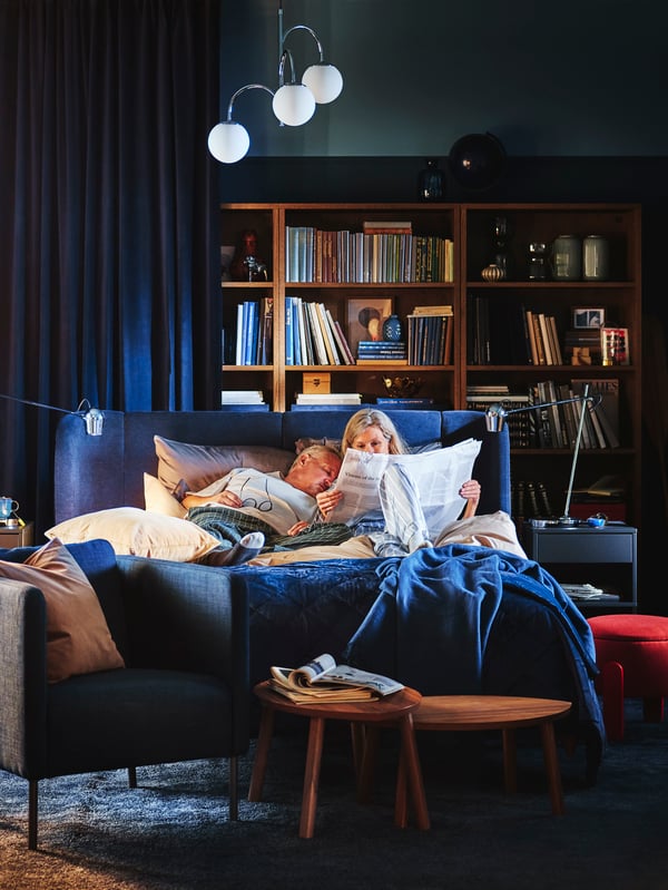 While a man sleeps, his wife reads a newspaper. LUKTJASMIN bedding and a MJUKPLISTER bedspread in dark blue are on the bed.