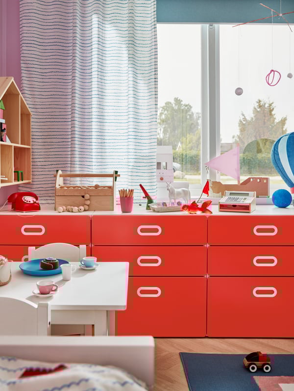 Three red STUVA/FRITIDS chests of 3 drawers stand in front of a window with various toys such as a telephone on top.