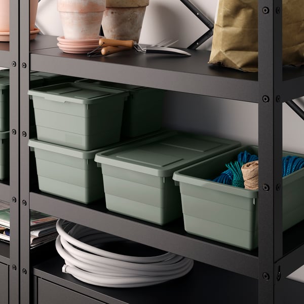 Two white SOCKERBIT boxes with lids are placed on a VAMDRUP textured rug. The SOCKERBIT box on the right is open and has a green INGRUN throw peeking out of it. A grey GURLI sofa sits behind the boxes.