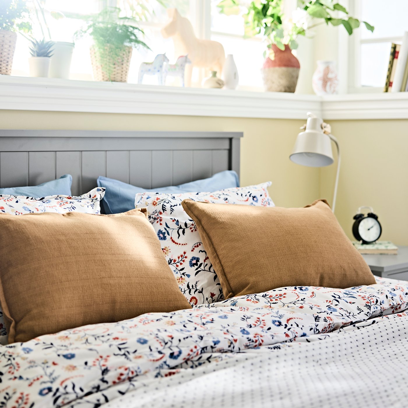 A bed with the BRUDKRUSBÄR duvet cover shown with large pillows, a grey headboard and a windowsill with plants above.