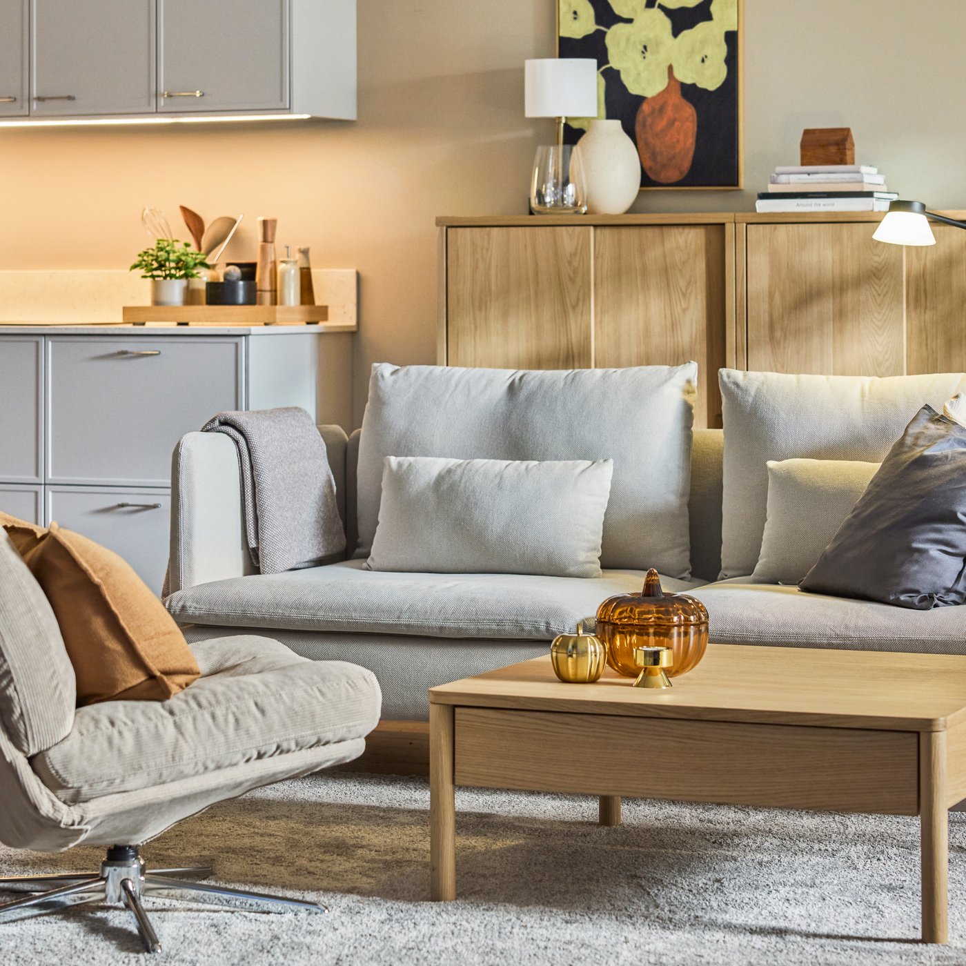 Cozy living room with sofa and armchair around wooden coffee table, soft textiles, warm lighting, kitchen area in background.