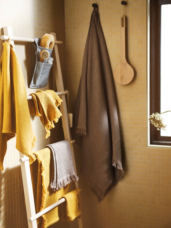 A bathroom with a VILTO towel stand in birch, towels, washcloths, shower brushes and two black hooks on the wall.