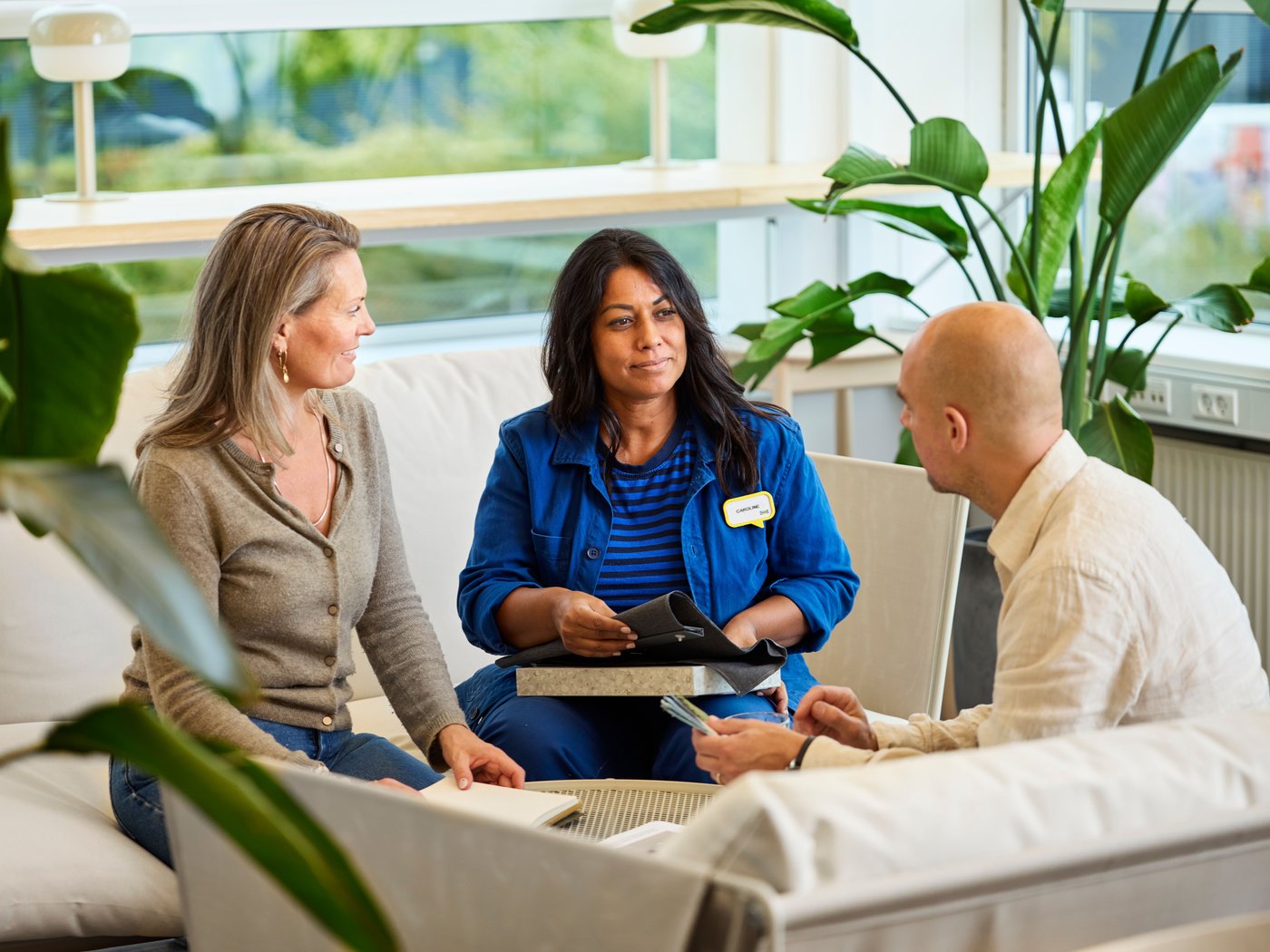 An IKEA interior designer with fabric samples in her hand sits on a sofa with two customers and advises them.