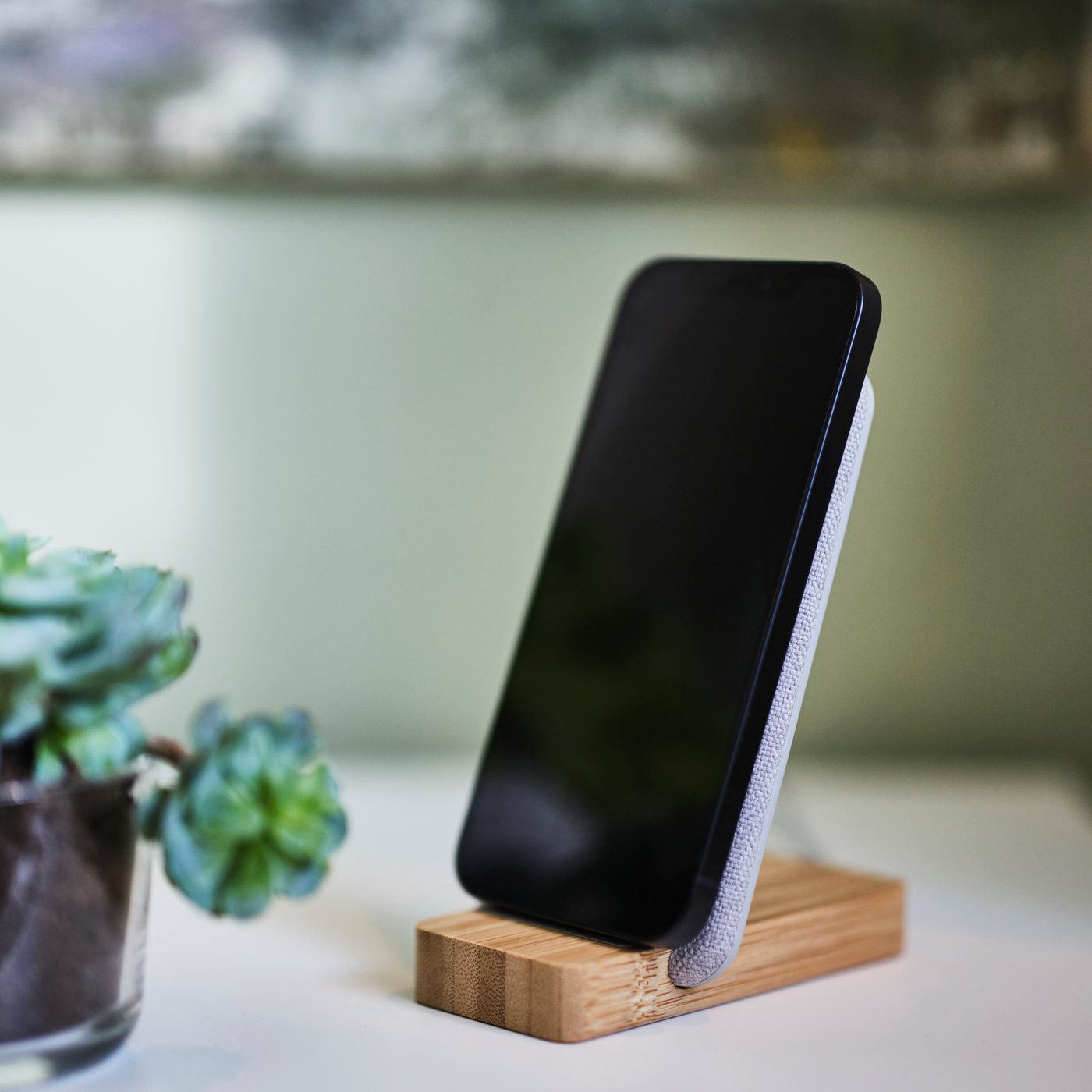 A bamboo NORDMÄRKE wireless charging stand charging a mobile phone on a white surface with a small, pot plant to the left.