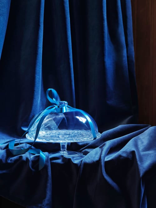 Close-up view of dining table with blue tablecloth, serving stand with lid made of glass, blue ribbon tied on the lid.