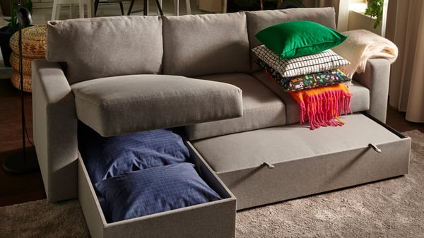 A grey BÅRSLÖV sofa bed with an open chaise longue part that offers storage for extra cushions and bedding. With a stack of an orange/bright pink KLIPPOXEL throw and green cushions.