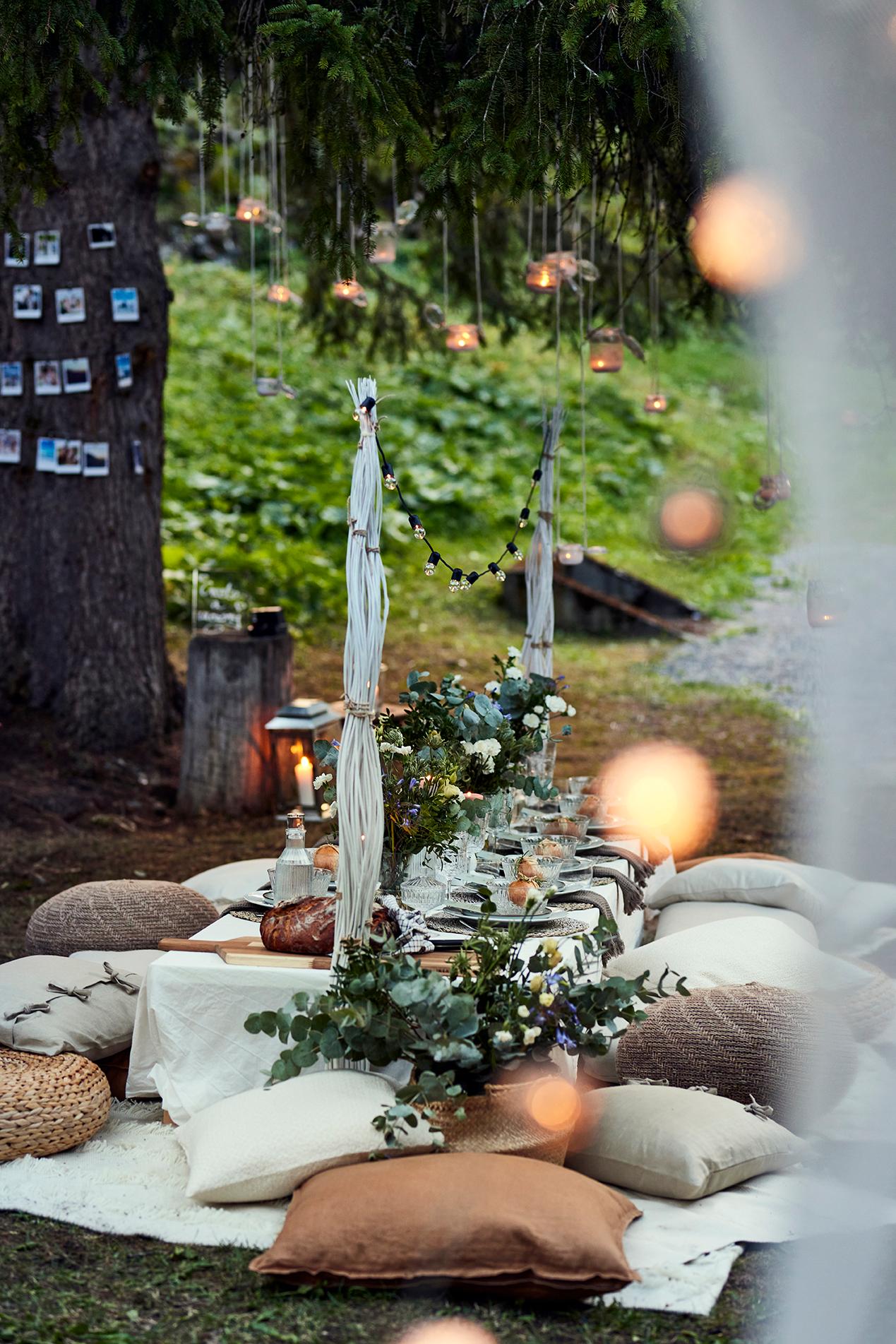 Outdoor-Tafel mit Kissen, Blumen und hängenden Windlichtern für ein stimmungsvolles Boho-Dinner im Grünen.