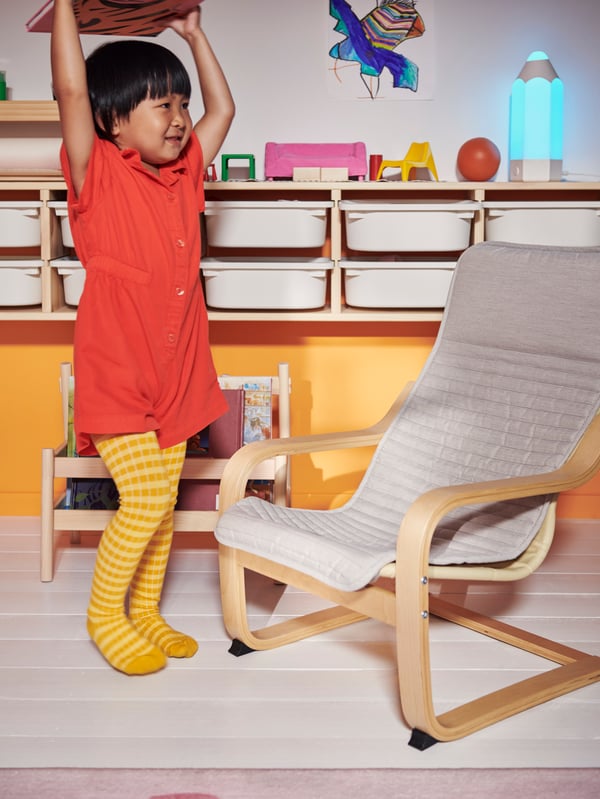 A child holding a book over their head in front of a FLISAT book display, and facing towards a light beige POÄNG armchair.