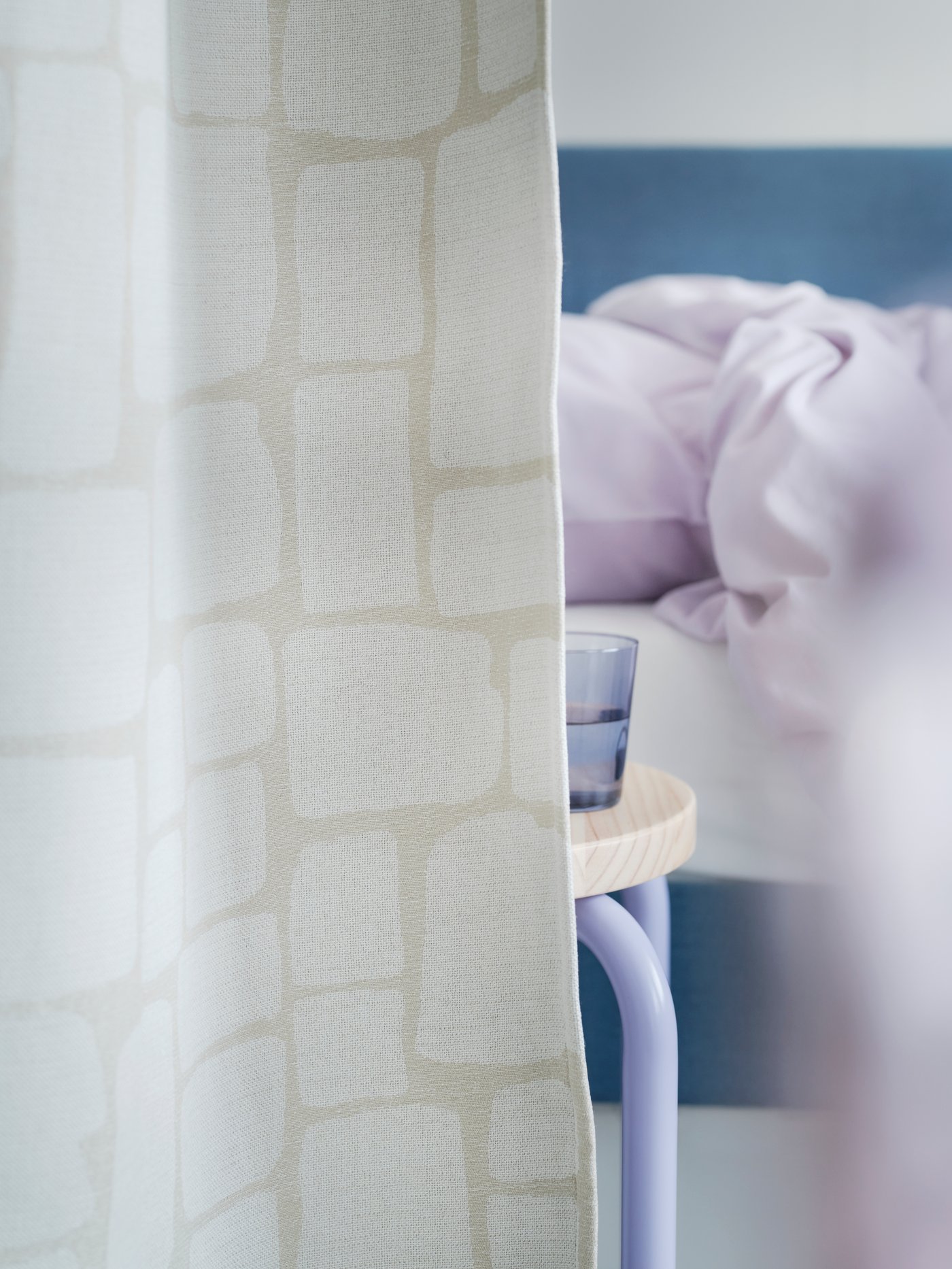 A close-up of a pair of LÖNNSTÄVMAL block-out curtains in beige as a room divider and a bed with purple bedlinen behind it.