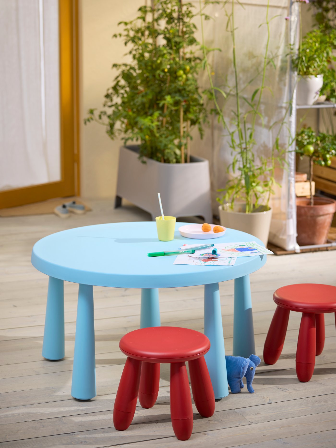 A light blue, round children’s table with pens and drawing-paper on it, and two red MAMMUT children’s stools next to it.