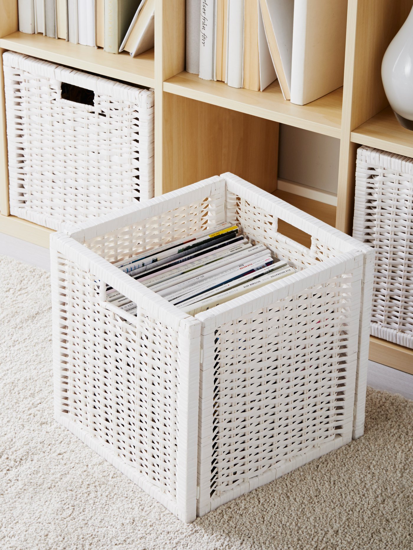 Scaffale KALLAX effetto rovere mordenzato bianco in soggiorno, con ripiani pieni di libri e cestini coordinati, e un cesto bianco in primo piano appoggiato sul tappeto chiaro.