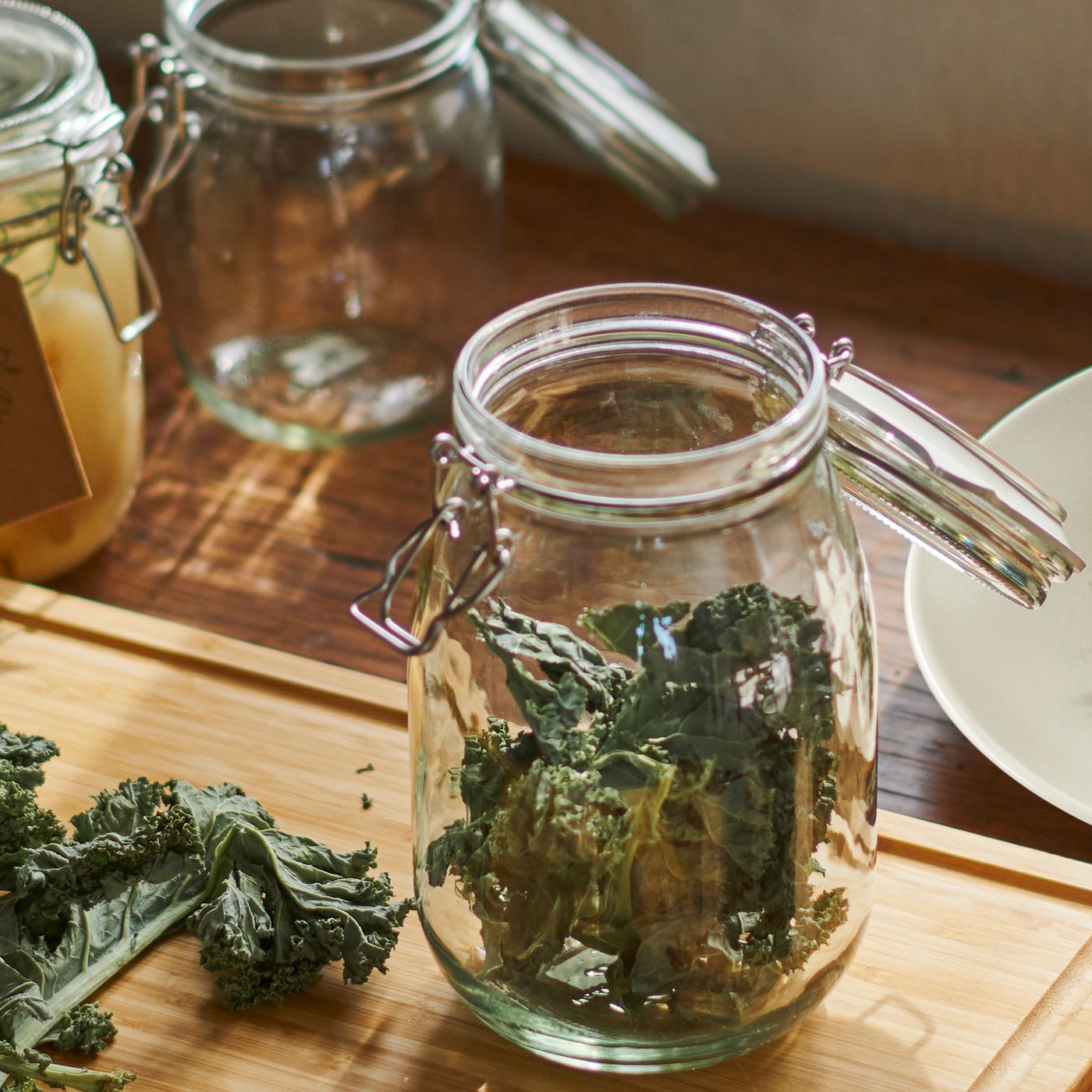 In a kitchen, a KORKEN jar with lid filled with kale chips is on a cutting board. Bits of kale are on the board near the jar.