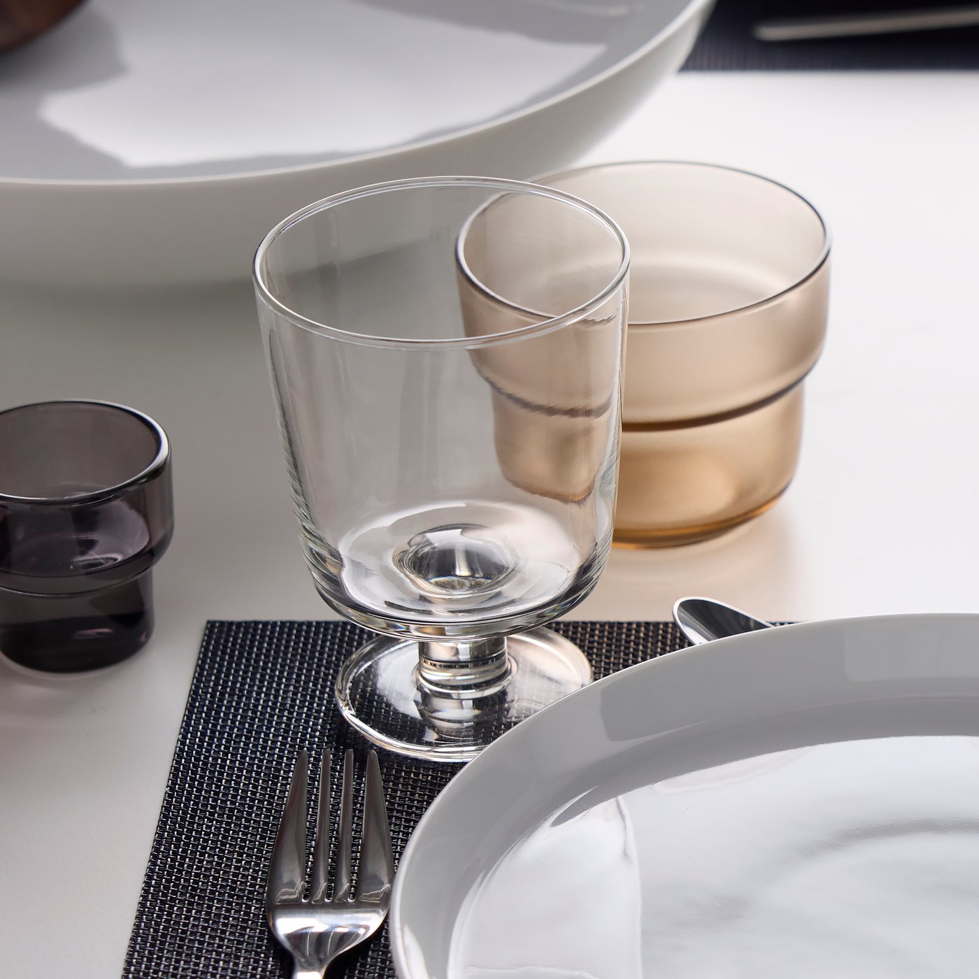 Part of a place setting at a table with an IKEA 365+ goblet, a light brown OLIVTRAST glass and an IKEA 365+ plate.