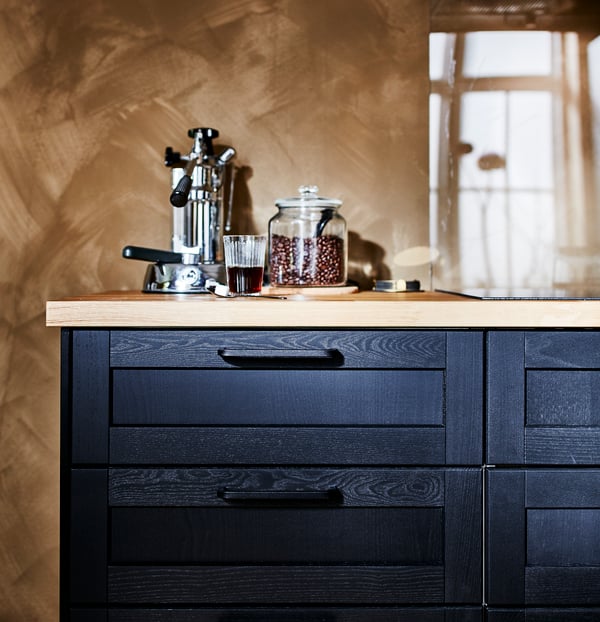 A black base cabinet with black drawer fronts and a wooden worktop. A coffee maker and a jar with coffee beans are on top.