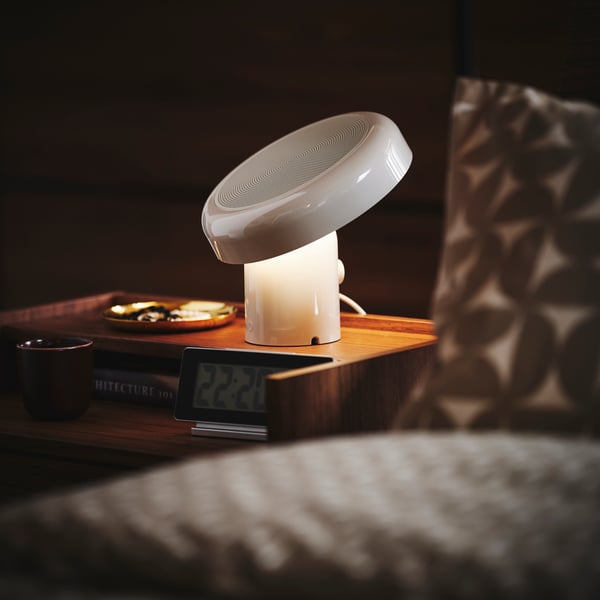 Close-up of a bedside table with BLOMPRAKT bluetooth speaker lamp, being the only light source in the otherwise dark room.