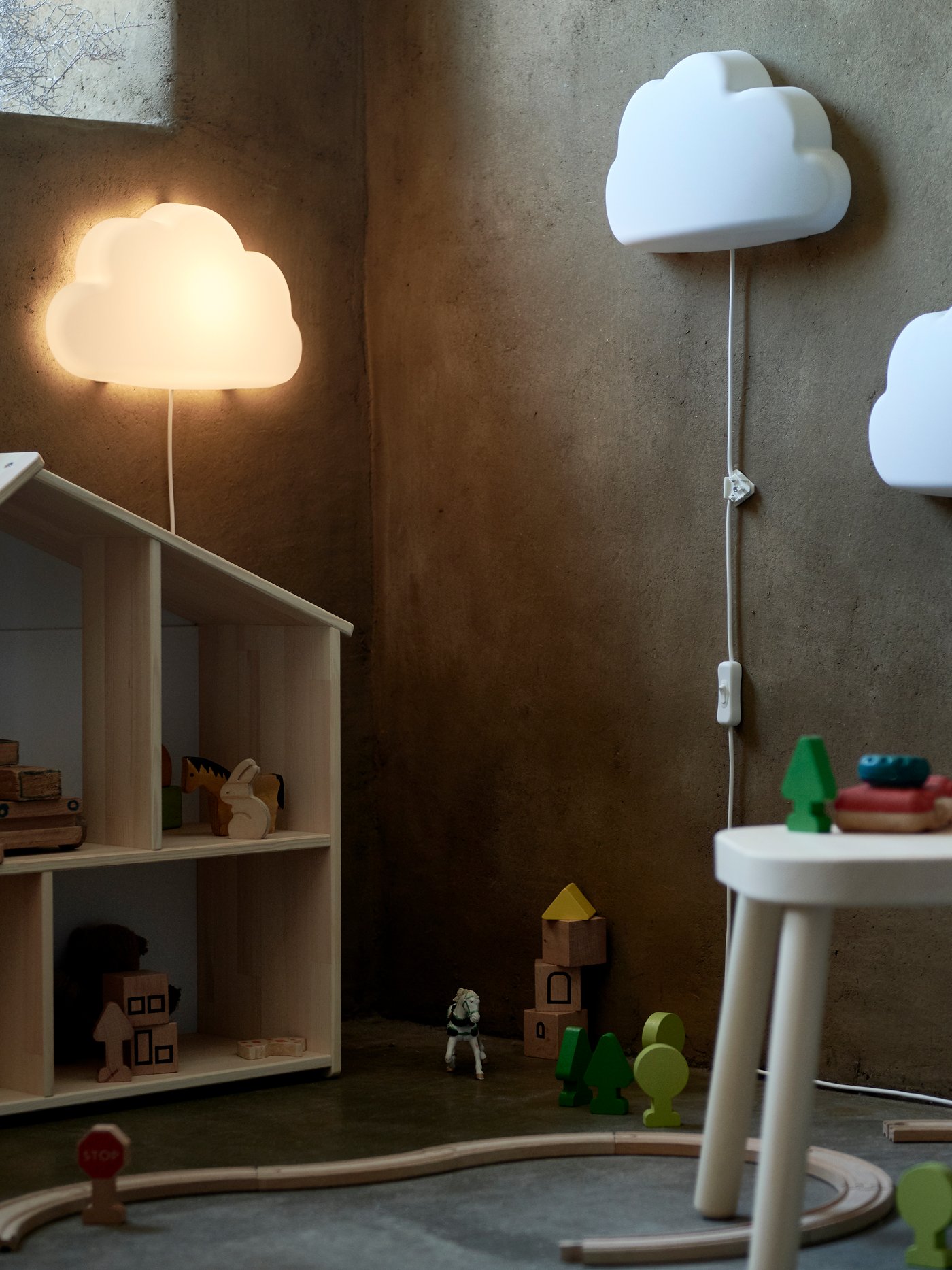 Children's room with two UPPLYST wall lamps shaped like clouds, one on and one off, dollhouse with wooden toys, round white stool with blocks and wooden train on the floor against a gray wall.
