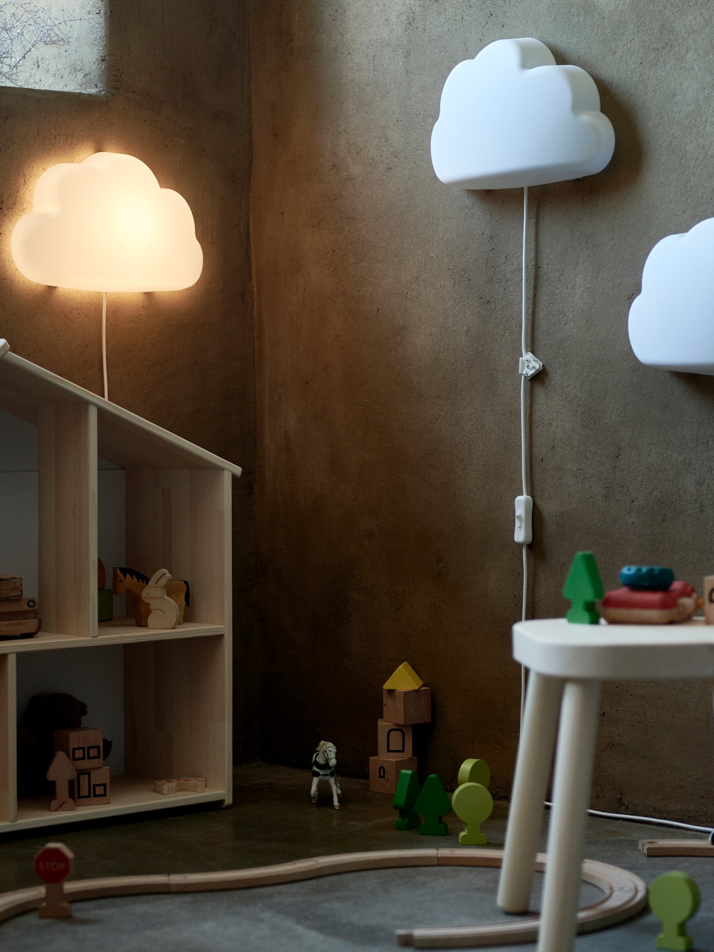 Barnrum med två UPPLYST vägglampor formade som moln, en tänd och en släckt, dockhus med träleksaker, rund vit pall med klossar och trätåg på golvet mot en grå vägg.
