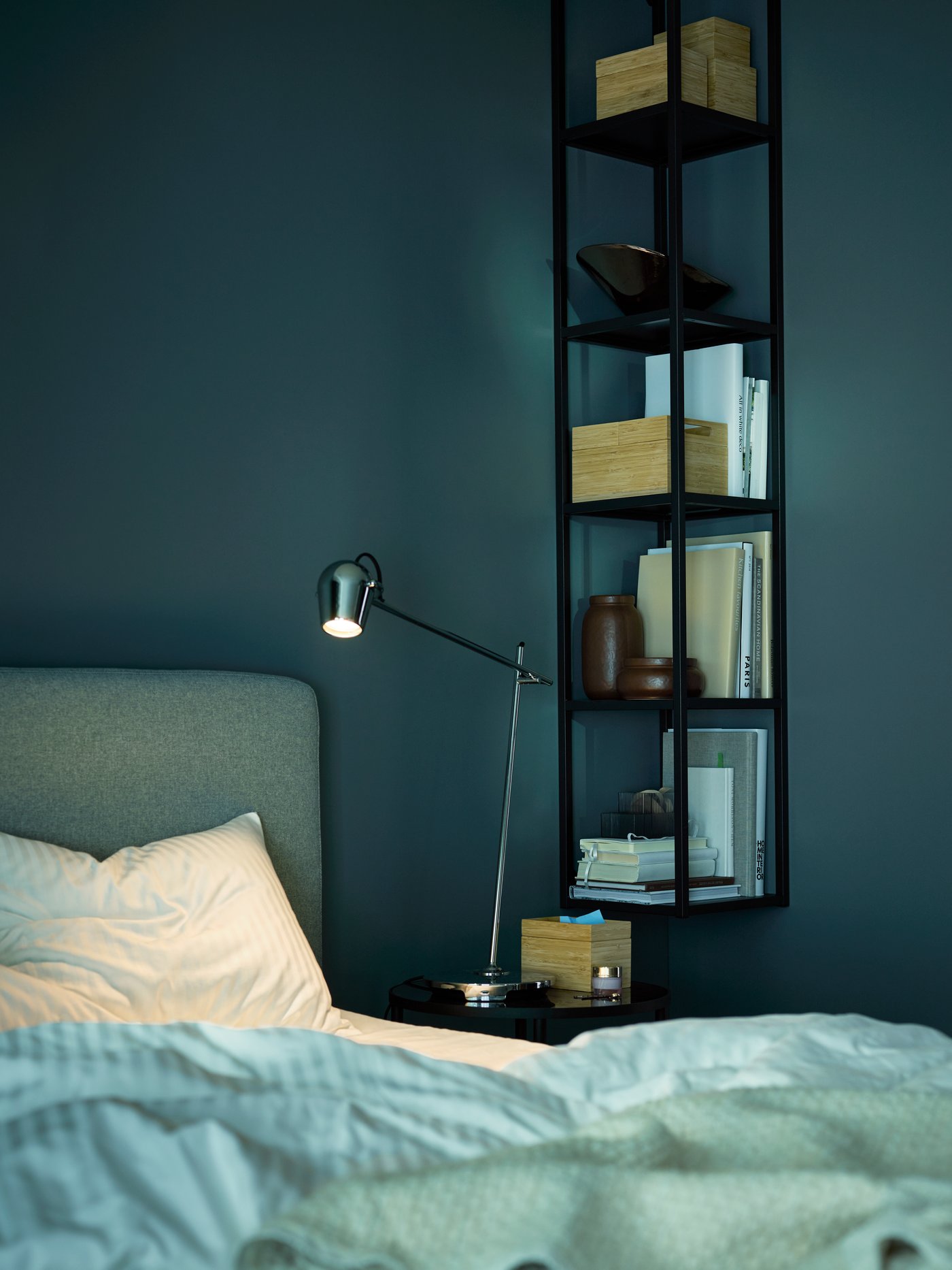 Hjørne af en seng med ENHET reoler på væggen og MODERMOLN arbejdslampe på natbordet. Værelset er mørkt, som om natten.