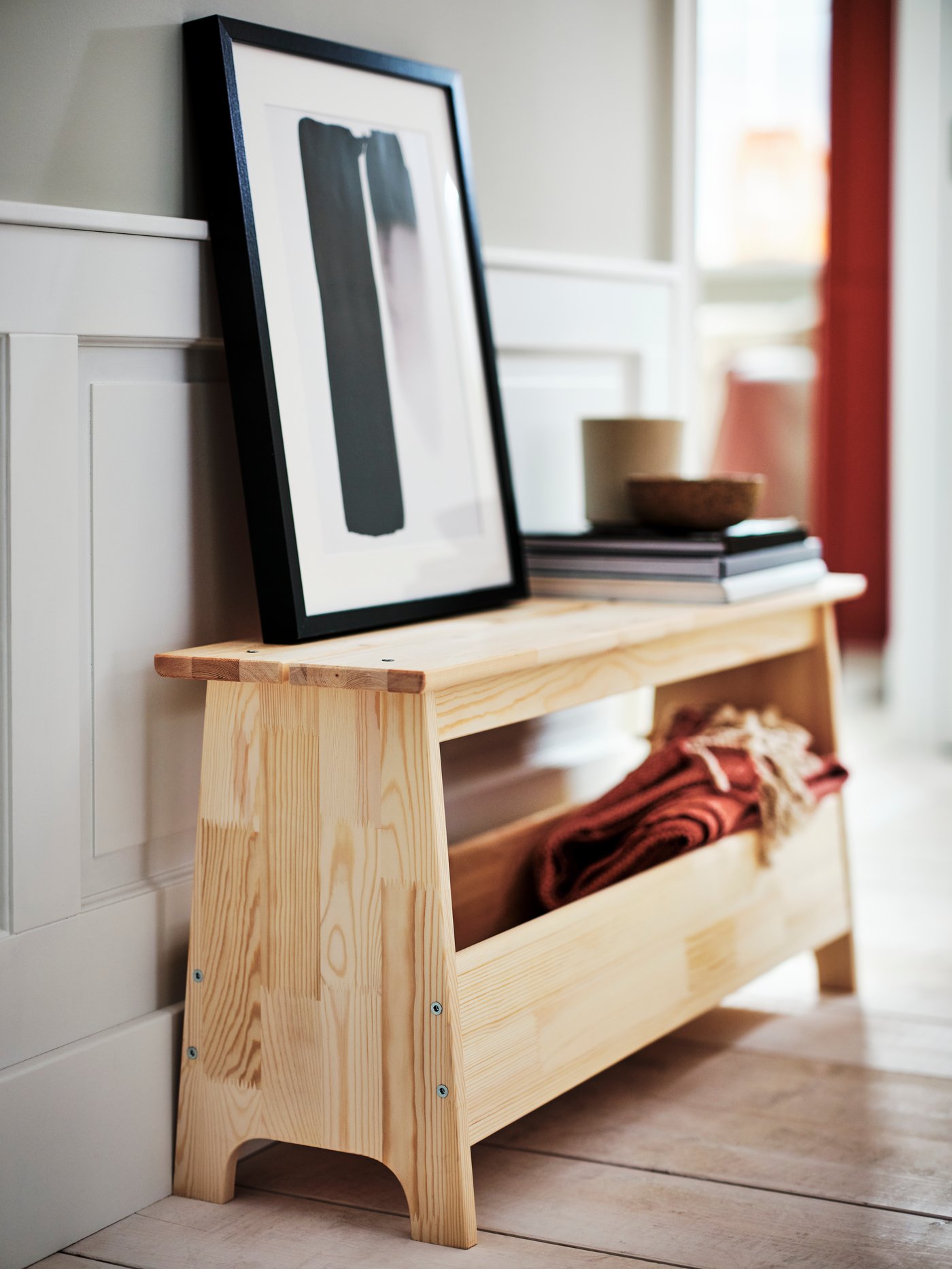 En BILD affisch i en svart RIBBA ram står lutad mot en vägg ovanpå en PERJOHAN bänk i furu med förvaring.