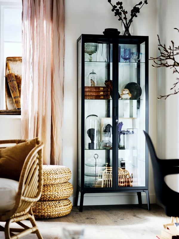 A black MILSBO cabinet with glass doors filled with an assortment of objects, in the corner of a bright living room.