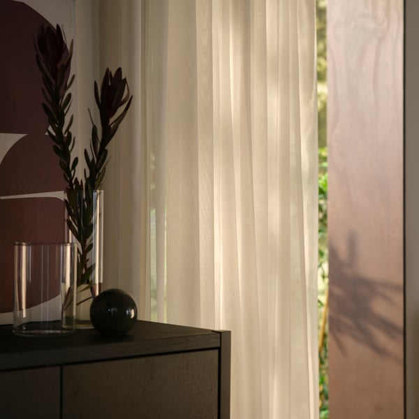 A living-room corner with a window covered by white STENFRÖ sheer curtains next to a dark-brown STOCKHOLM 2025 cabinet.