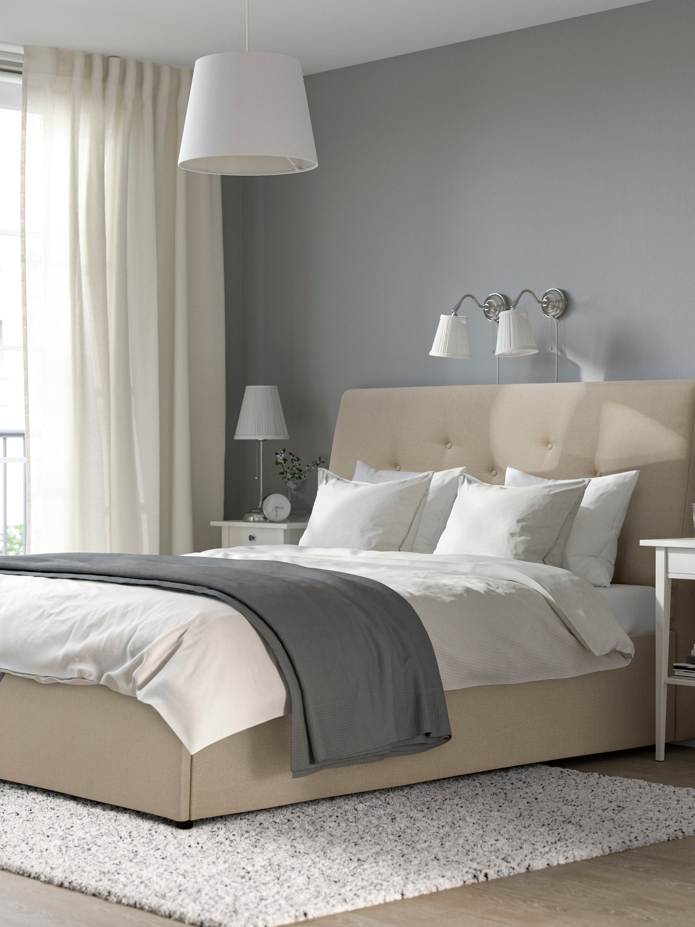 Letto contenitore IDANÄS rivestito in tessuto beige Naggen in una camera da letto luminosa, con biancheria bianca, coperta grigia, comodini coordinati e grande finestra con tende chiare.