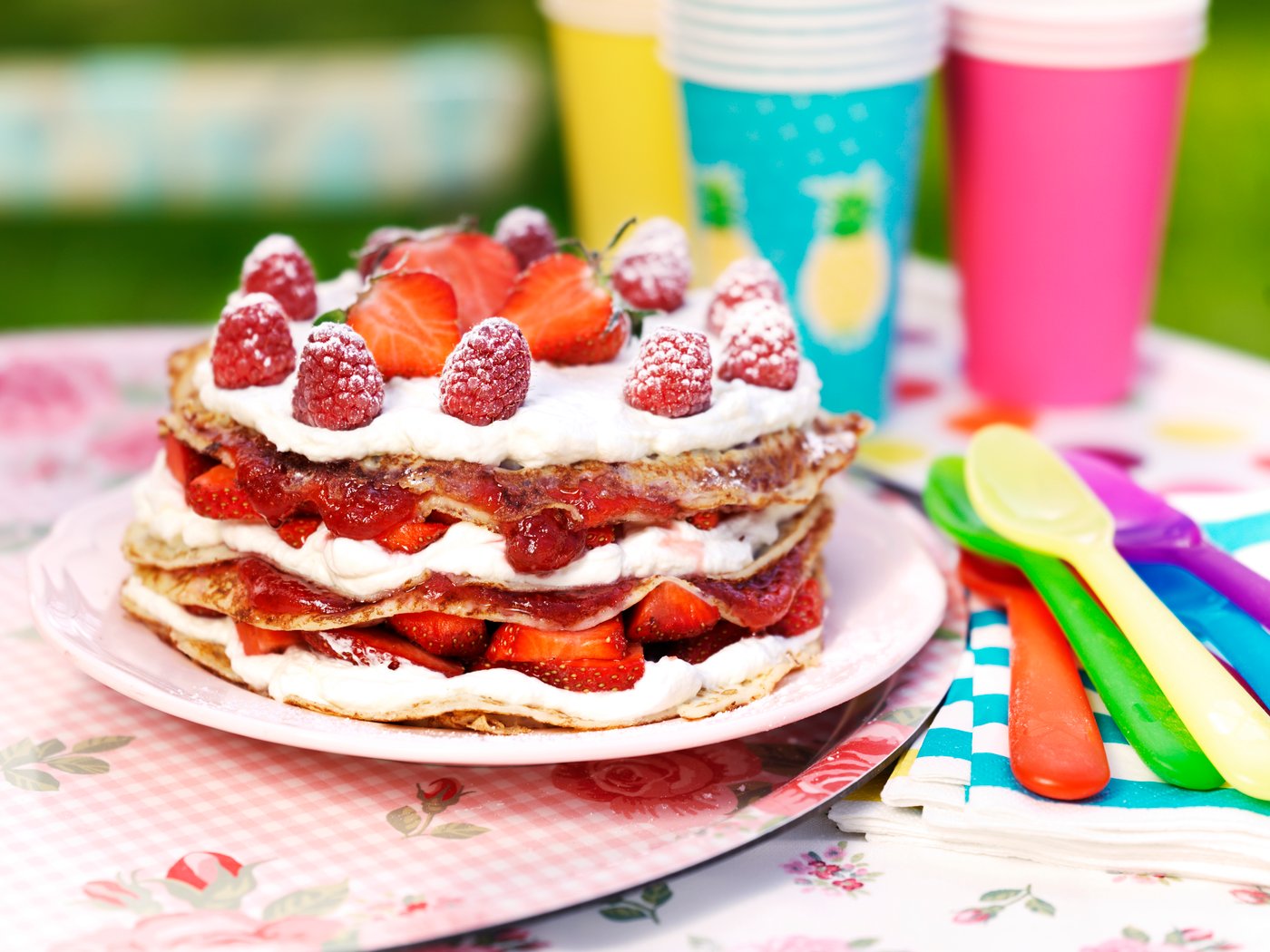 Kihiline pannkoogitort, mille vahel on vahukoor, moos ja värsked maasikad ning mille peal on vaarikad ja terved maasikad. Tort on asetatud lillelise mustriga laudlinale õues.