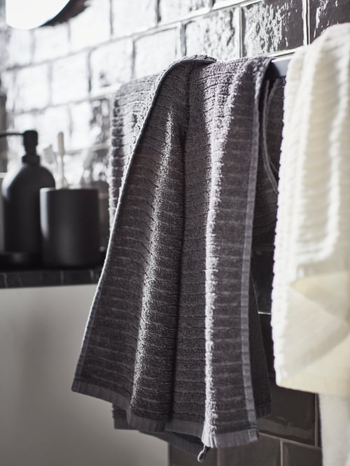 A dark grey VAGJSON bath towel with striped design and a white towel are on a towel rail next to a grey soap dispenser.
