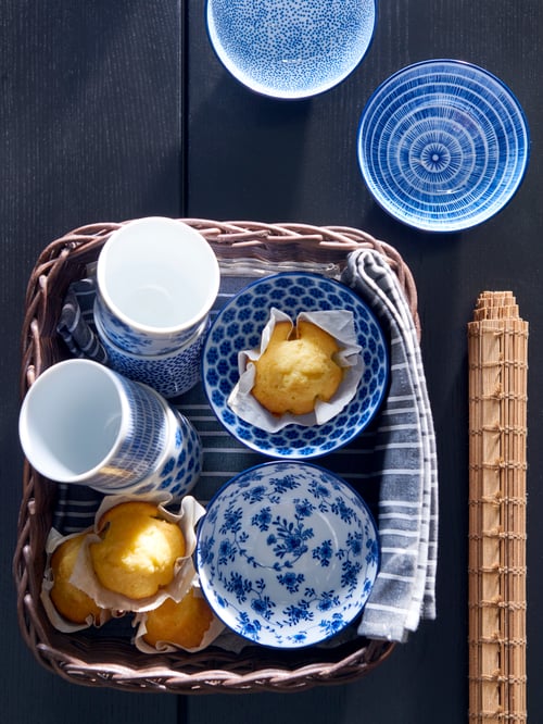 Table seen from above with a basket of four bowls and four cups with white and blue patterns, and a place mat next to it.