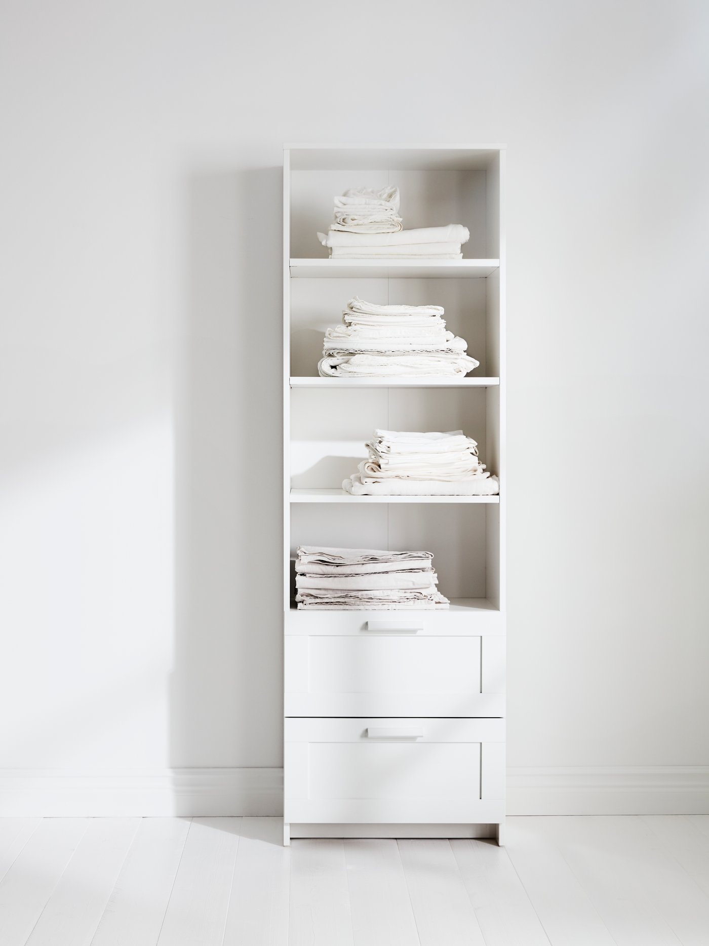 Una librería BRIMNES blanca con dos cajones y cuatro estantes. Junto a una pared blanca, hay pilas de sábanas blancas dobladas.