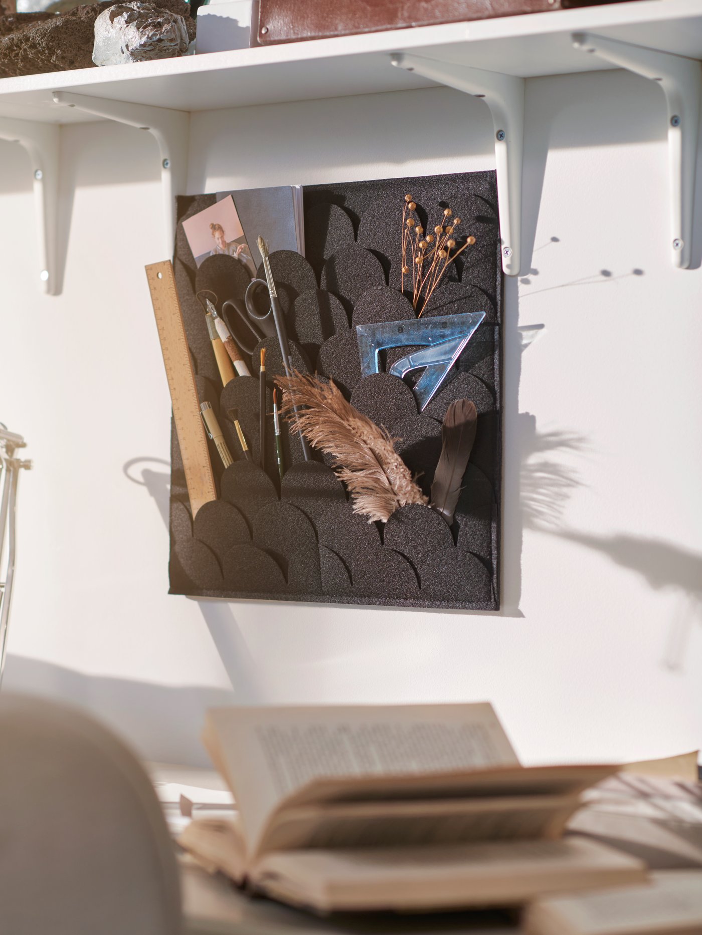 A PLUGGLAND memo board hangs on the wall underneath a shelf. It holds pens, scissors, dried flowers and feathers.