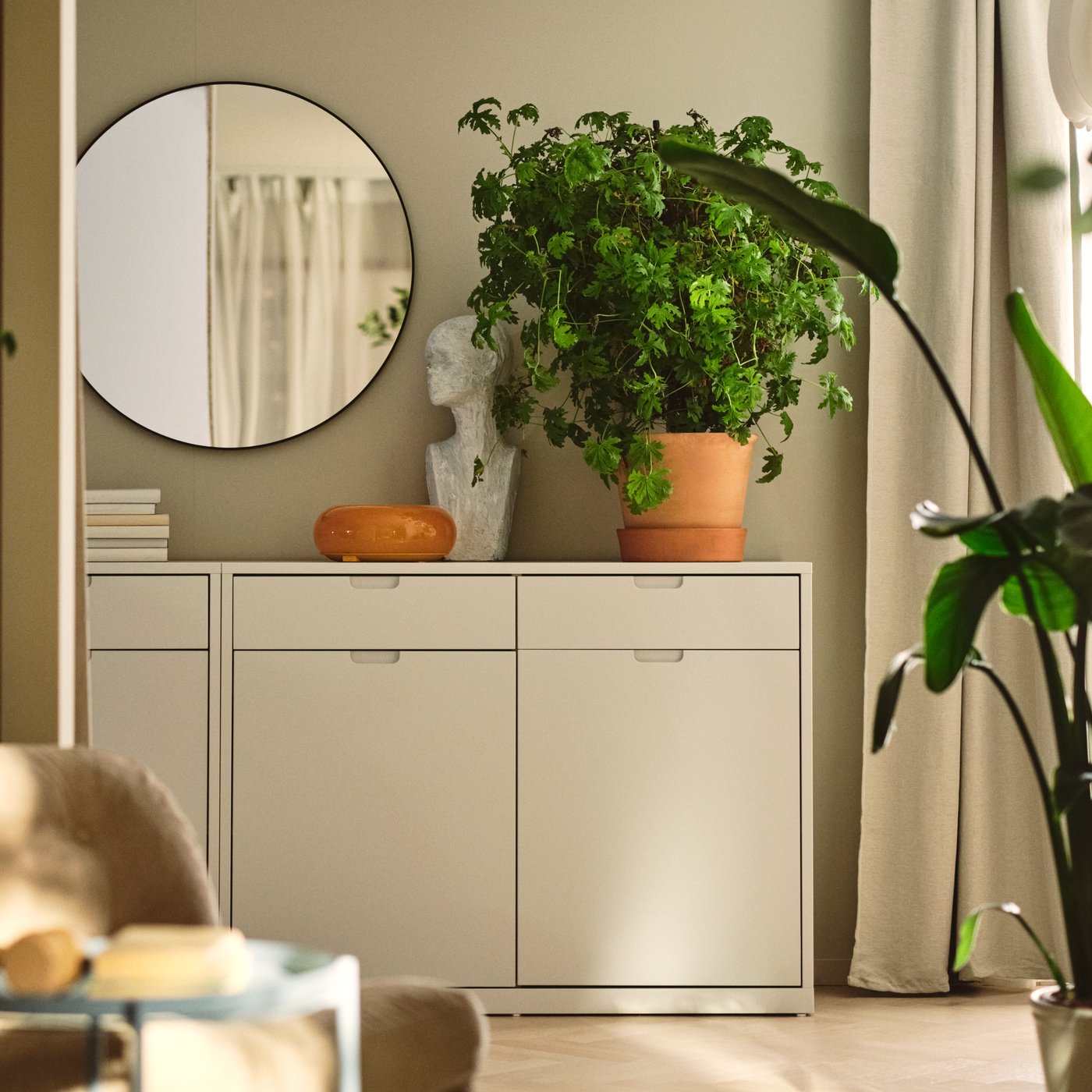 A living room with an off-white TONSTAD sideboard and a large round mirror, beside a lush green plant.