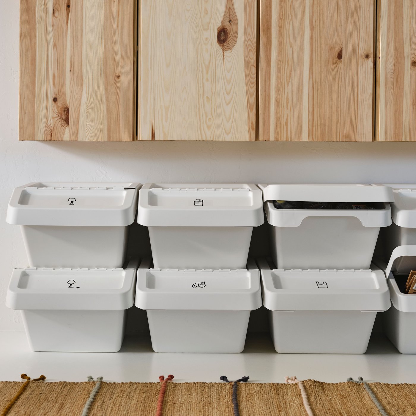 Es ist eine Reihe mit übereinandergestapelten weißen SORTERA Abfalleimern mit Deckel unter einer Reihe Schränke in Kiefer zu sehen. Davor liegt ein Teppich.