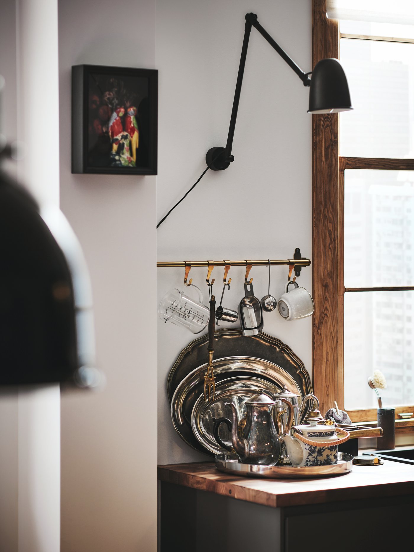 Cocina con una lámpara de trabajo de pared flexible de acero en color negro sobre una encimera y sobre un riel de pared con utensilios y tazas.