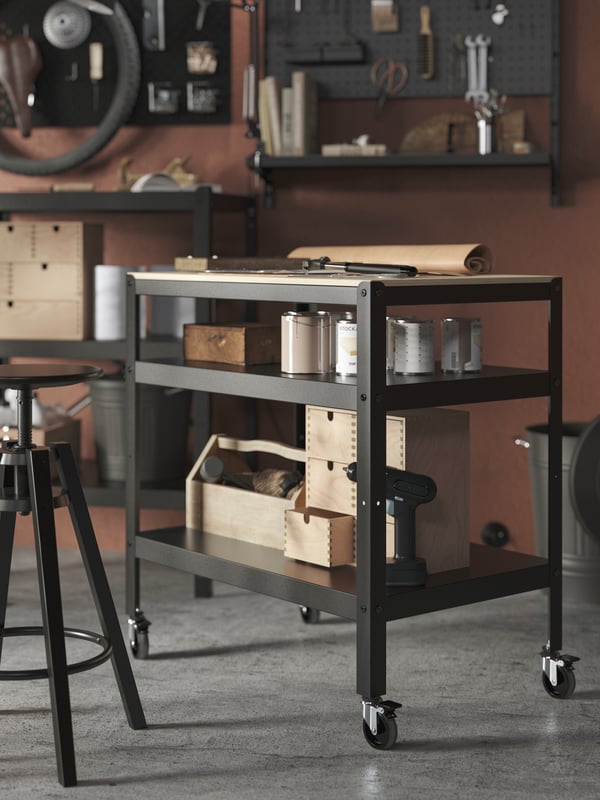 An organised garage interior with a DALFRED bar stool next to a black-and-pine-plywood BROR trolley holding tools.