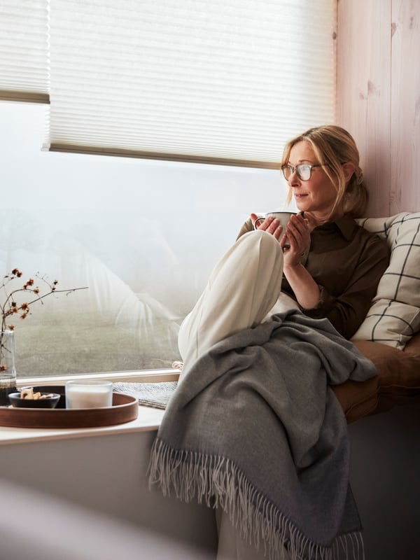 Eine Person ruht sich auf ein paar Kissen aus, die auf einer Fensterbank liegen. Zwei weisse PRAKTLYSING Wabenjalousien sind hier halb geschlossen vor einem Fenster zu sehen.