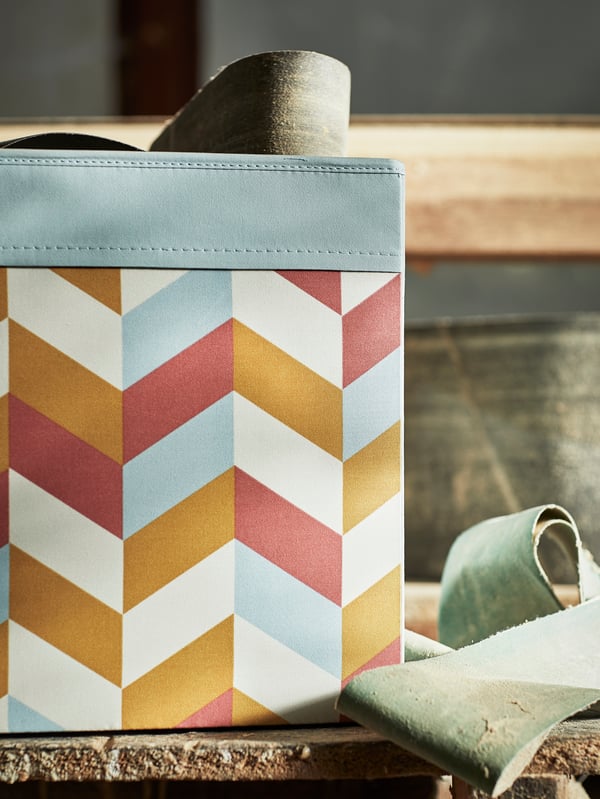 Uma caixa DRÖNA com um padrão multicor sobre uma prateleira antiga, com algumas tiras pequenas de lixa colorida.