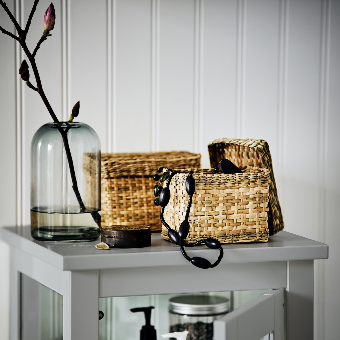 Two seagrass LURPASSA boxes by a vase on top of a HEMNES high cabinet. One box is open with a necklace hanging out.