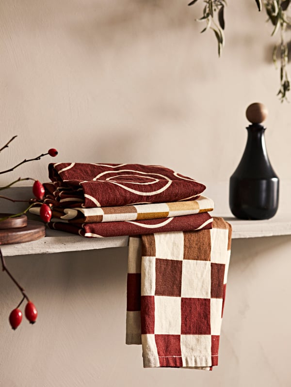 Folded KRÖSAMOS tea towels together with a KRÖSAMOS black bottle on a wooden shelf inside a kitchen.