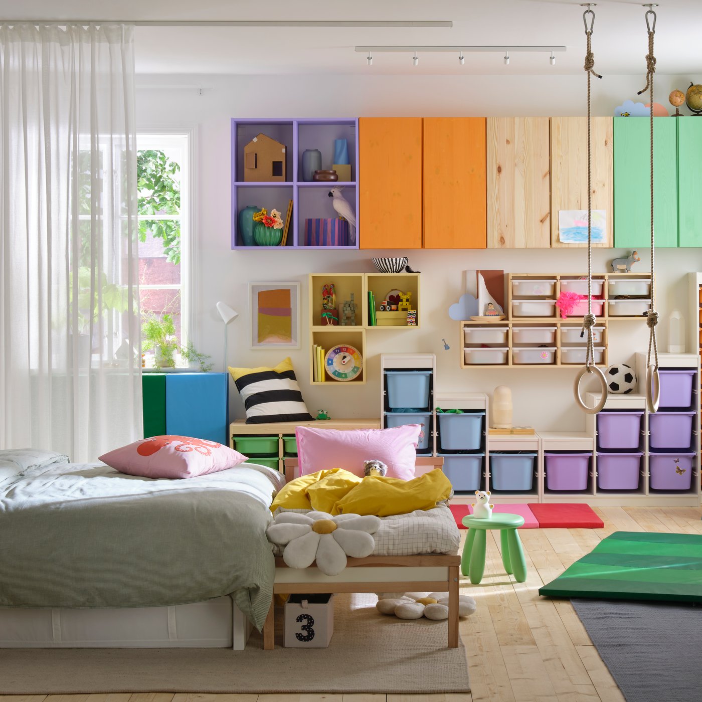 Ett färgglatt barnrum med förvaringslådor, hyllor och skåp i olika färger, en bäddad säng och en lekplats med gymnastikringar på golvet.