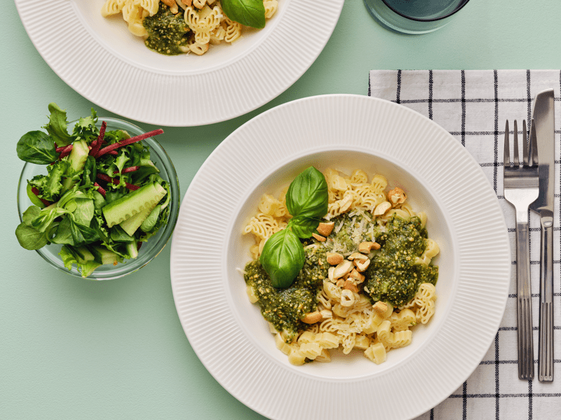 Due piatti fondi bianchi, contenenti pasta BÄSTISAR, pesto e anacardi tritati, su una superficie verde chiaro.