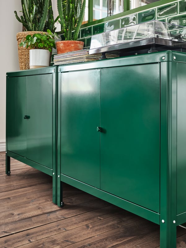 Two green cabinets stand by a green-tiled wall. Plants, a pile of records and a record player are on the top surfaces.