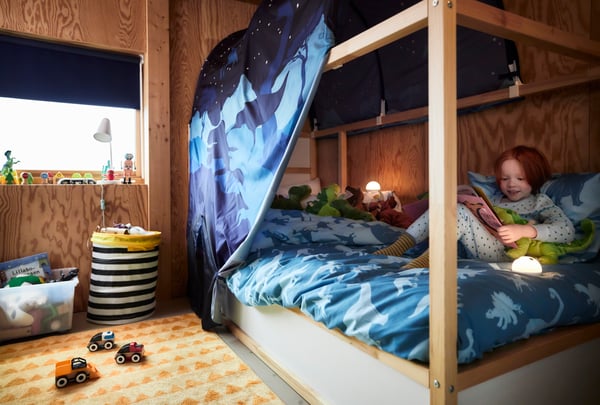 A white KURA reversible bed in a children’s room with a dinosaur themed blue tent and blue quilt cover, and a child.